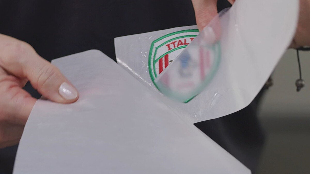 [Translate to Hongkong (Englisch):] embroiderer holding a badge with heat seal film to fif the badge onto a garment